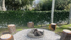 Ein kreisförmiger Sitzbereich mit Feuerstelle, umgeben von weißen Kieselsteinen und Holzstühlen mit karierten Kissen.