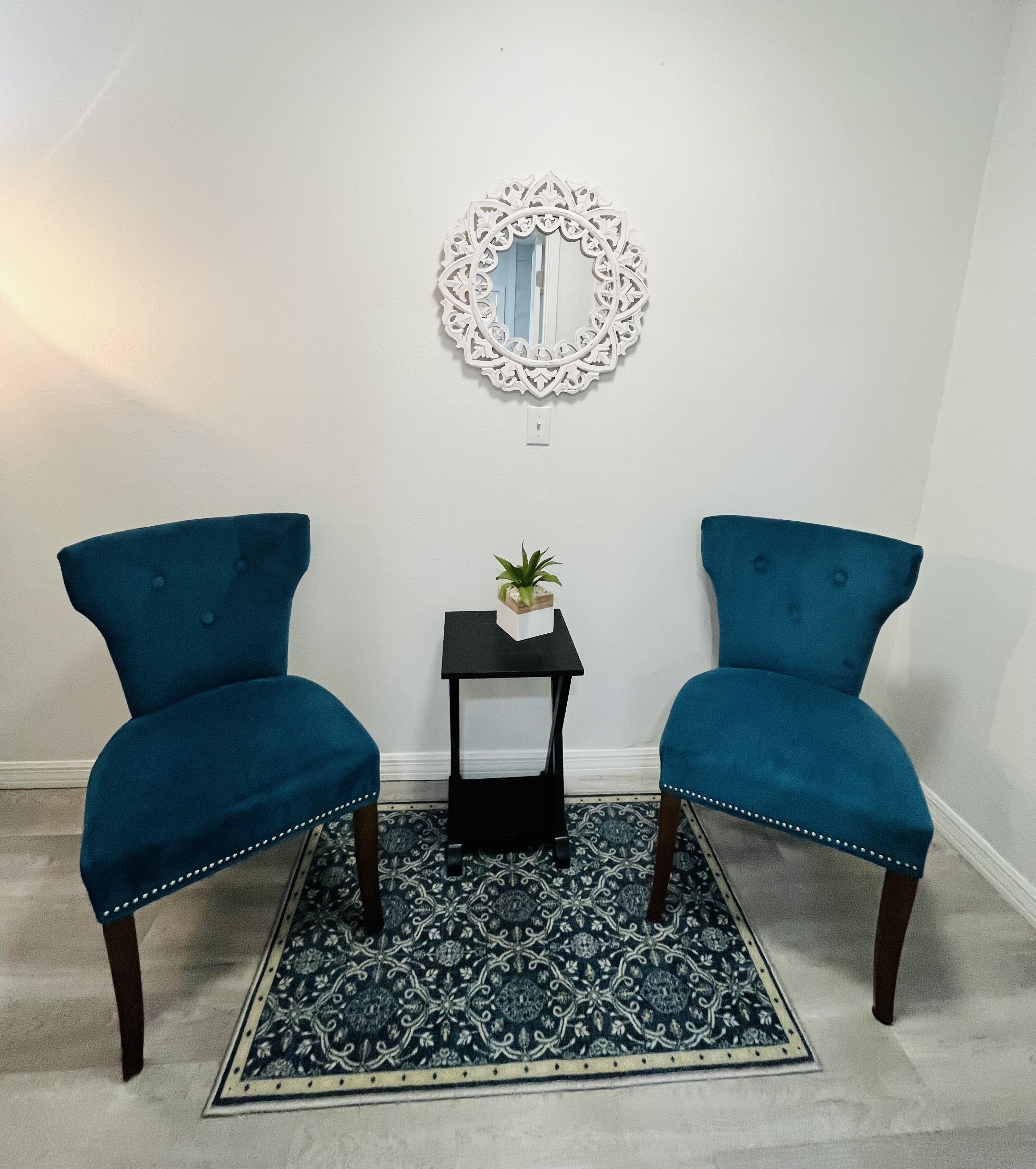 Two blue armchairs, a small black side table with a plant, and a decorative mirror on a patterned rug in a room with white walls.