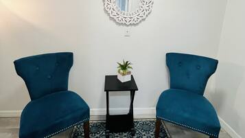 Two blue armchairs, a small black side table with a plant, and a decorative mirror on a patterned rug in a room with white walls.