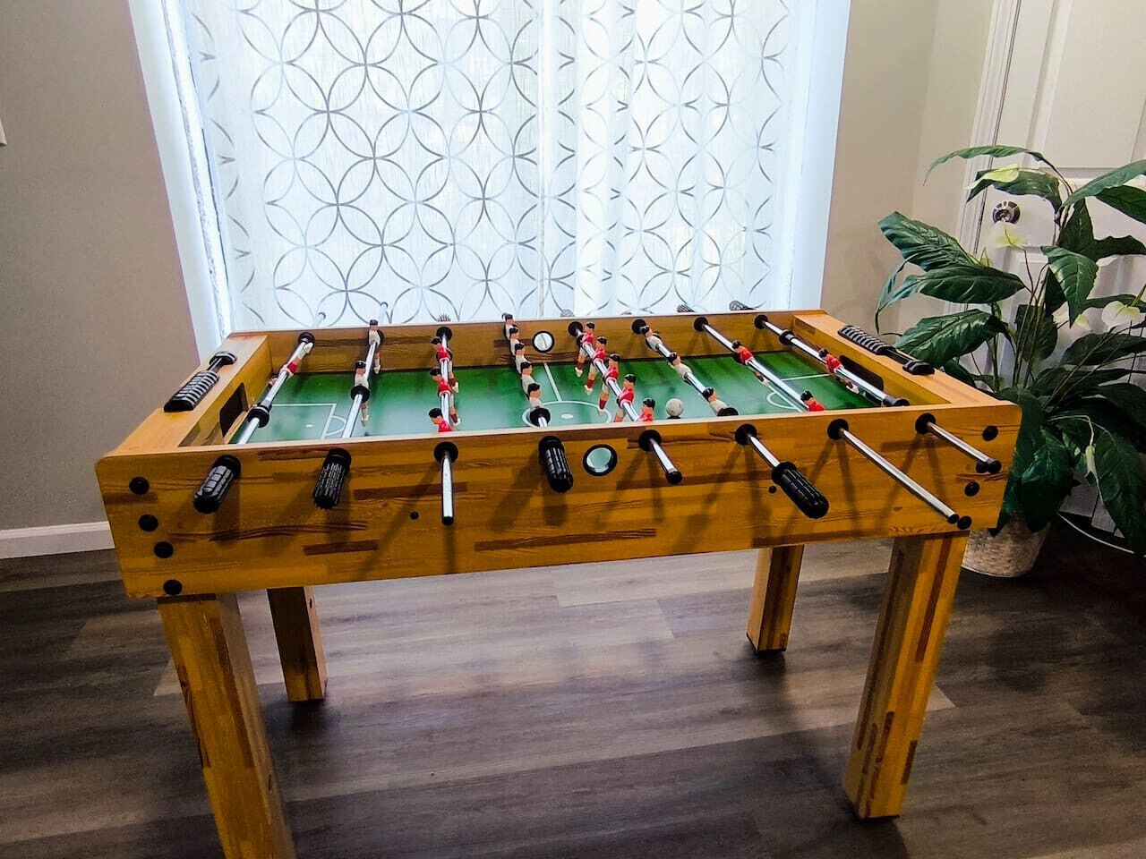A wooden foosball table with a green playing surface, positioned in a room with a patterned window and a potted plant.