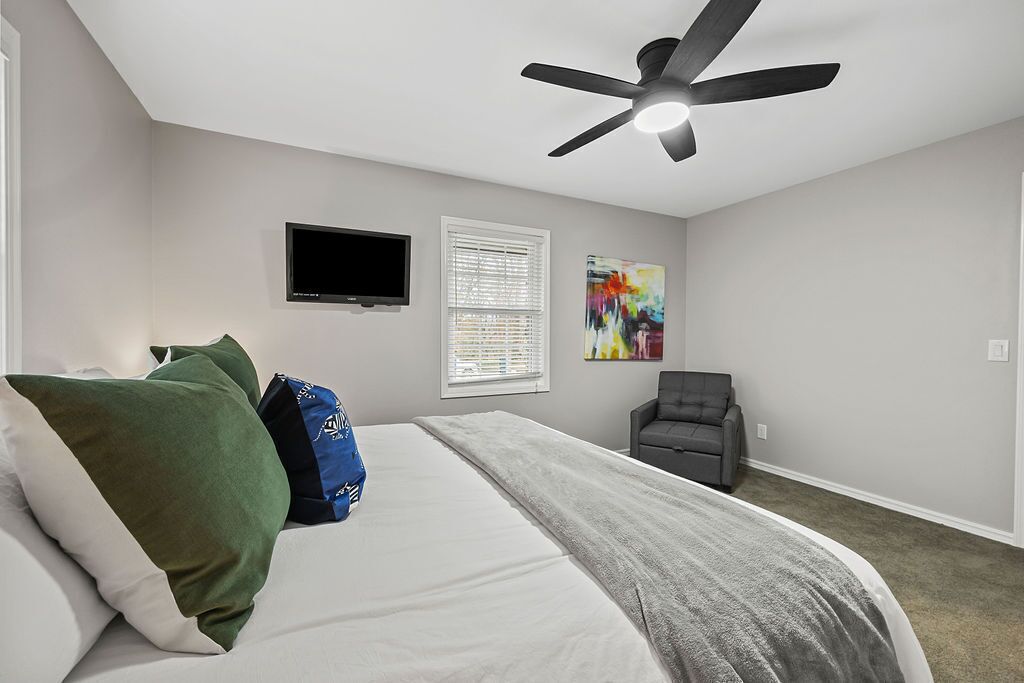 Ein Schlafzimmer mit Bett, Kissen, Fernseher, Deckenventilator, Stuhl und Wandbild.
