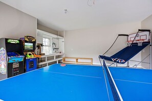 Una habitación con una mesa de ping pong azul, un aro de baloncesto pequeño y máquinas de arcade.