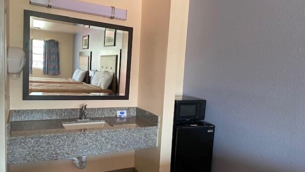 A hotel bathroom with a granite countertop sink, a mirror, a black refrigerator, and a visible shower area. - Texas Inn (Seguin)
