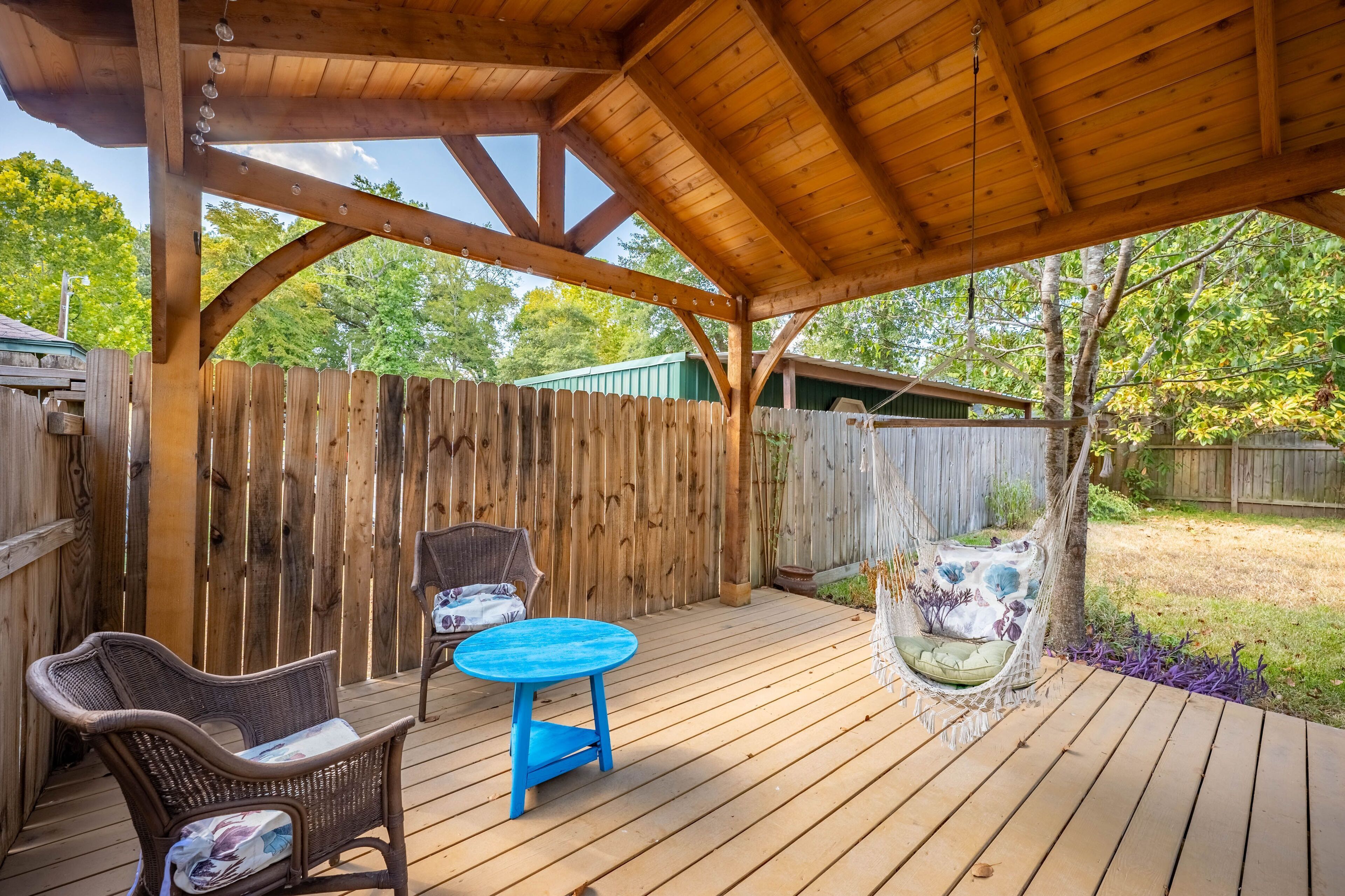 A wooden deck with a covered area, two wicker chairs, a blue table, and a hanging chair.