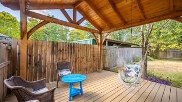 A wooden deck with a covered area, two wicker chairs, a blue table, and a hanging chair.