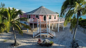 Een roze strandhuis met balkon, omringd door palmbomen en een zandstrand.