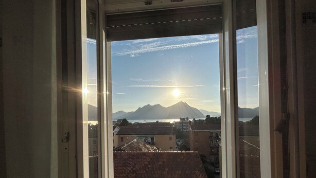 A view from a window with a clear sky, mountains, and a body of water in the distance. - LAKEHOUSE ENJOY (Pallanza)