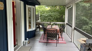 Een overdekt terras met een tafel en stoelen, een rode deur en uitzicht op groen.