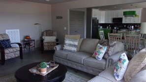 Sala de estar com sofá, poltronas, uma mesa de centro com uma planta e uma cozinha ao fundo.