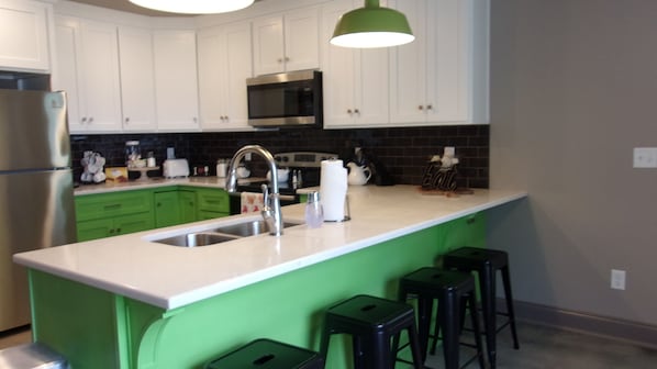 Uma cozinha com armários brancos, geladeira de aço inoxidável, micro-ondas e uma ilha verde com banquetas.