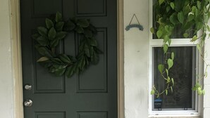 A green door with a wreath, a window with a hanging plant, and a 'Welcome' sign above it.