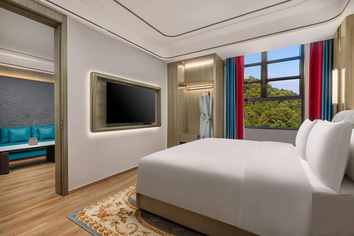 Quarto de hotel moderno com uma cama grande, televisão, área de estar com sofá e vista para o verde pela janela.