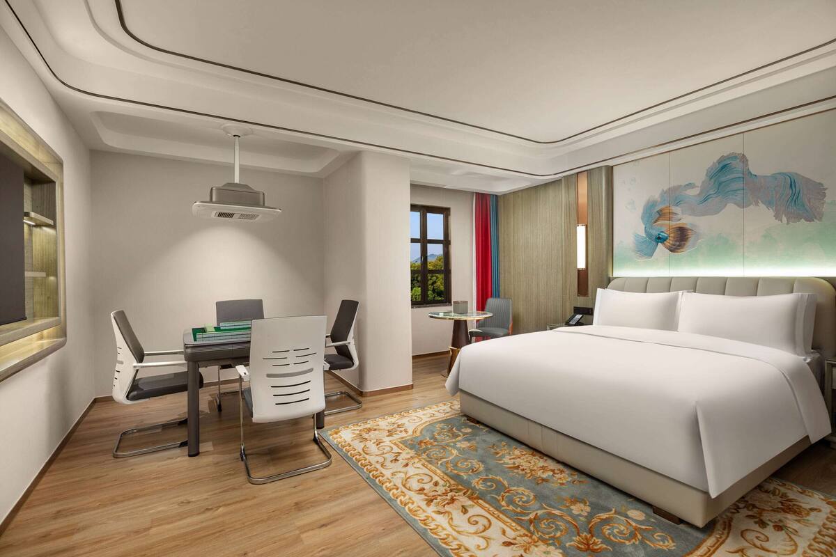 Quarto de hotel com cama, escrivaninha, cadeiras e uma janela grande.