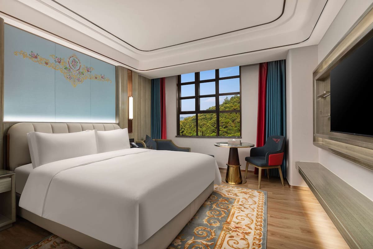Quarto de hotel com uma cama grande, uma escrivaninha com cadeira, uma TV na parede e uma janela com vista para o verde.