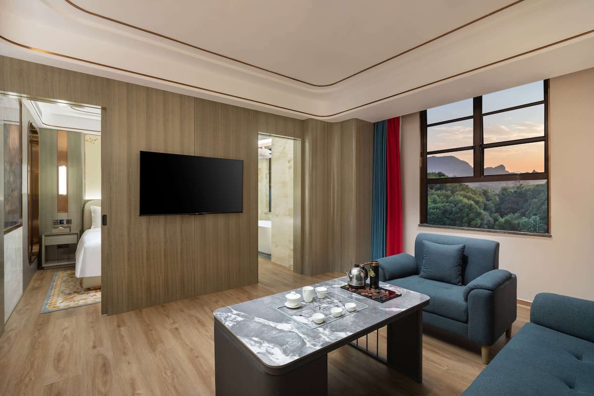 Quarto de hotel moderno com TV de tela plana, mesinha de centro de mármore, sofá e uma grande janela com vista para o exterior.