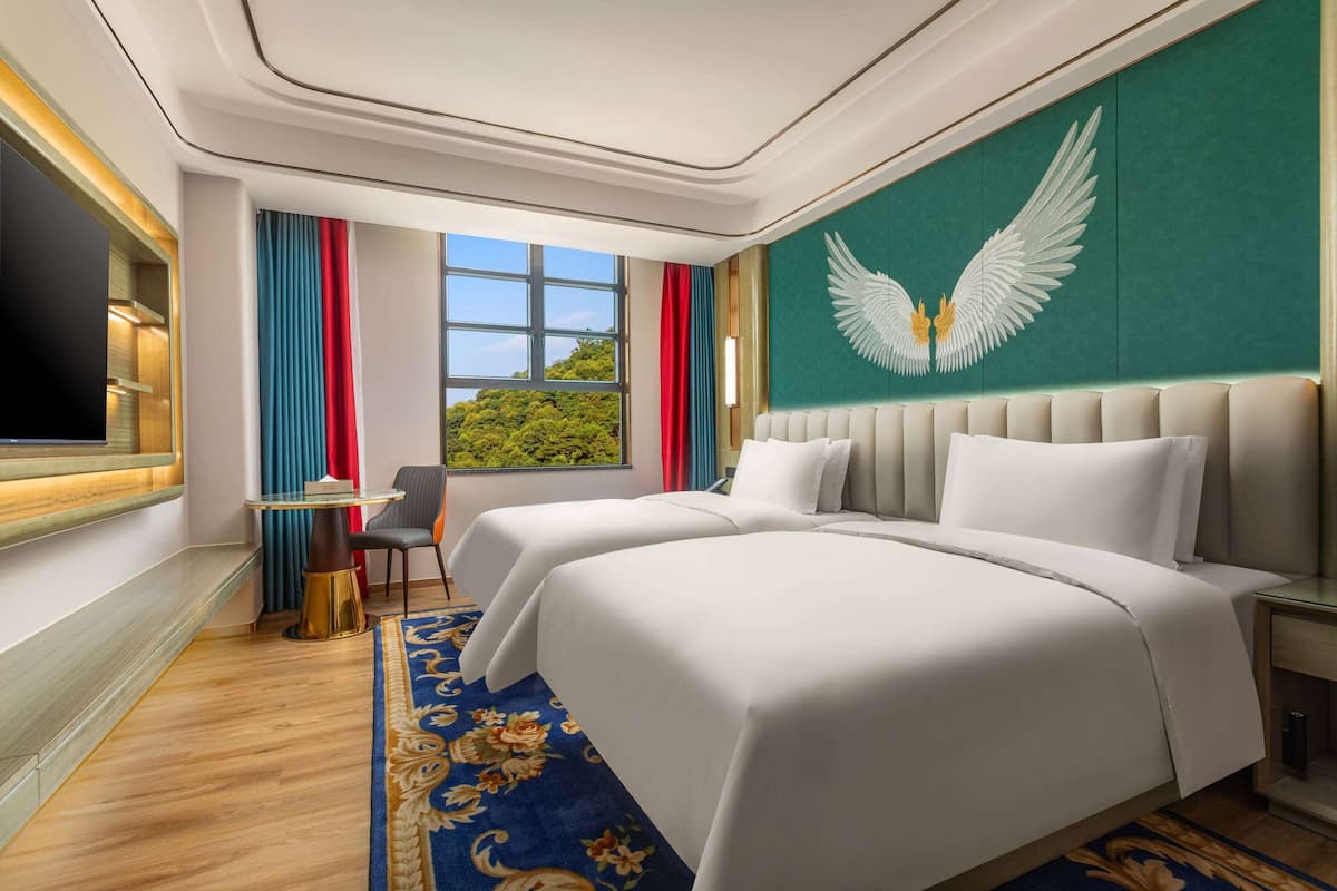 Quarto de hotel com duas camas, uma escrivaninha, uma cadeira, televisão e uma janela grande com vista para áreas verdes.