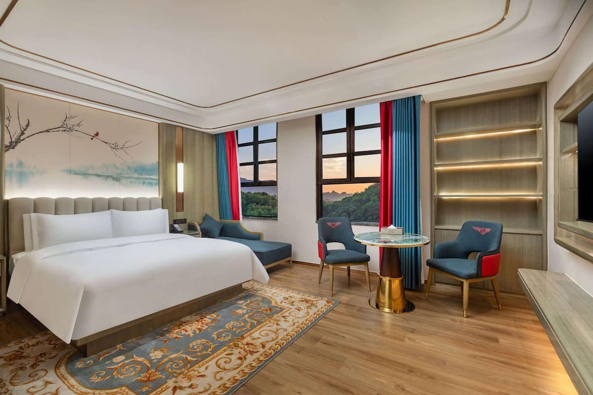 Quarto de hotel com uma cama grande, duas cadeiras, uma mesa pequena e vista para uma paisagem montanhosa pelas janelas.