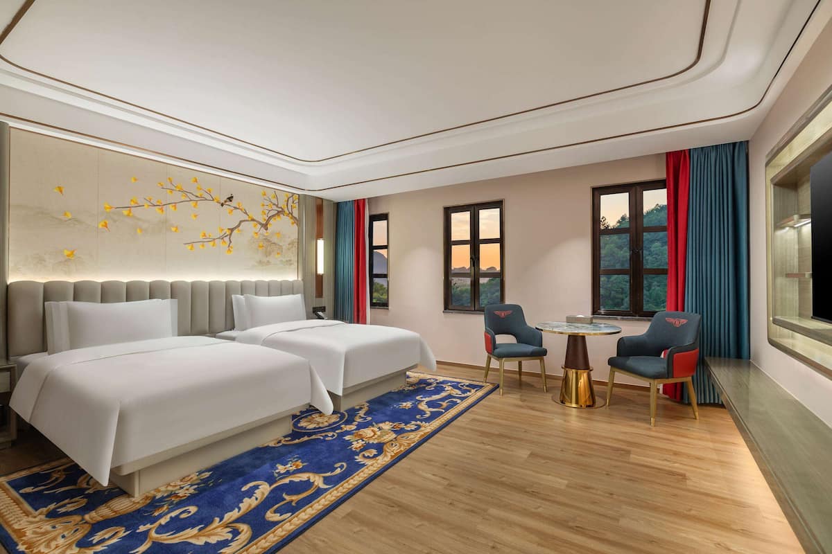 Quarto de hotel com duas camas, uma mesa pequena, duas cadeiras, parede decorativa e janelas amplas.