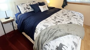 Ein Schlafzimmer mit Holzboden, einem Bett mit gemustertem Überwurf, einem Nachttisch mit Lampe und einem Fenster mit Vorhängen.