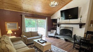 Ein geräumiges Wohnzimmer mit Holzbalkendecke, Kamin, Wand-Fernseher und einem großen Fenster mit Blick ins Freie.