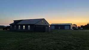 Una casa grande con garaje y un cobertizo más pequeño, rodeada de un campo cubierto de césped.