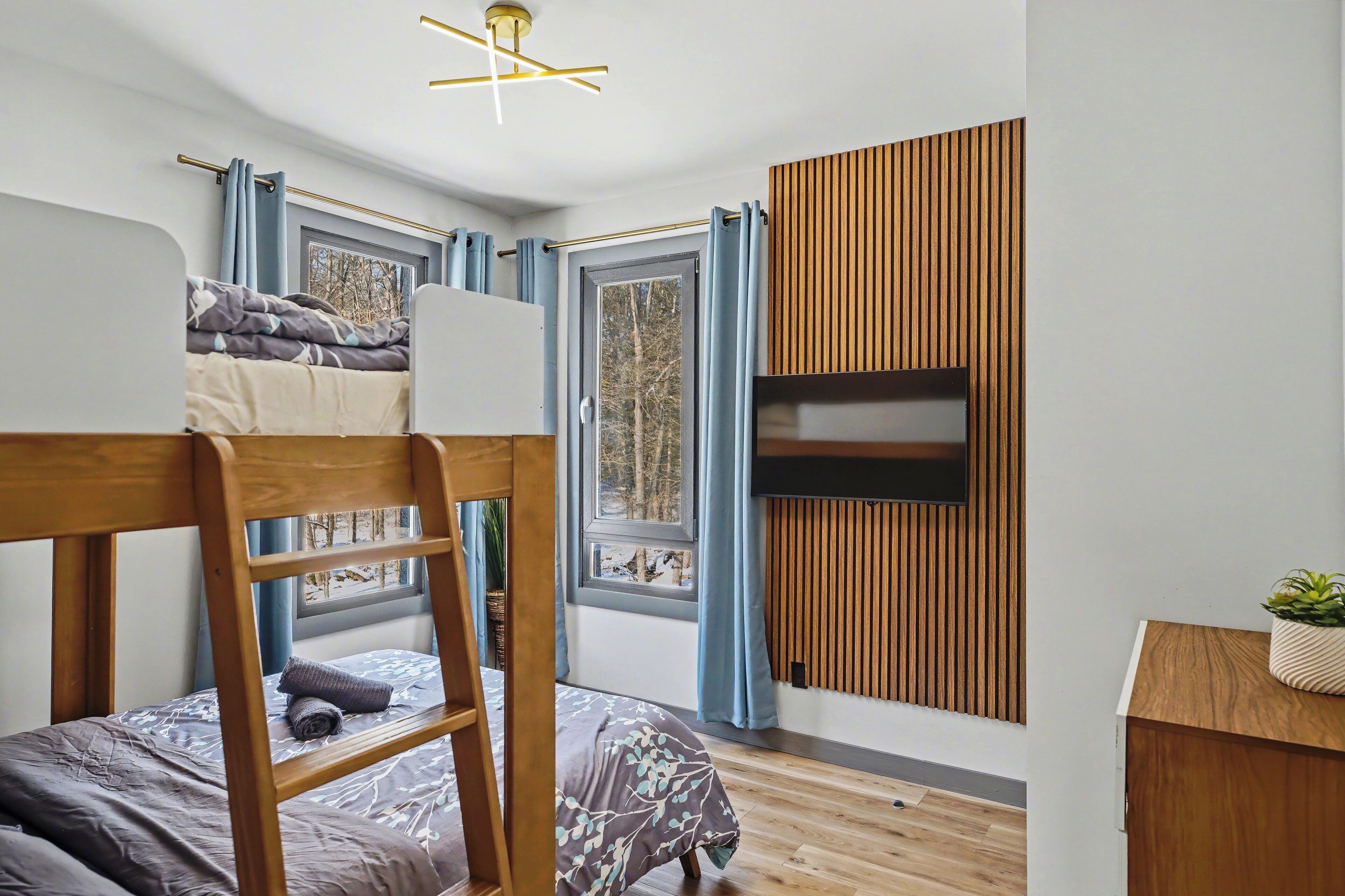 Une chambre moderne avec un lit superposé, un bureau, un téléviseur fixé sur un mur recouvert de panneaux en bois et une fenêtre avec des rideaux.