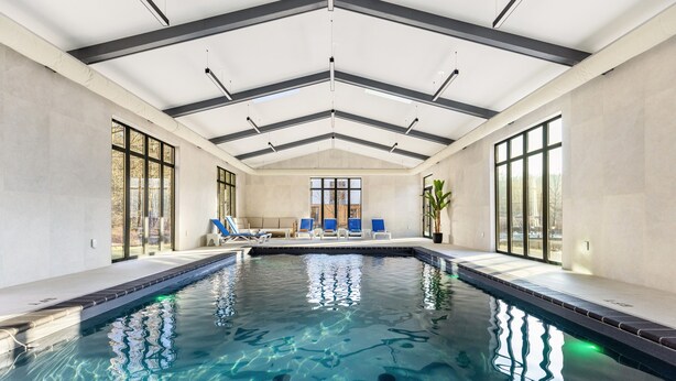 An indoor swimming pool with clear water, a ceiling with black beams, and a seating area with blue chairs.