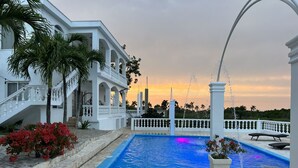 Un área de piscina con un edificio blanco, palmeras y un atardecer.