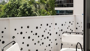 A balcony with two white chairs and a small table, a perforated wall, and a view of a modern building and greenery. - City Center Piantini W/ Pool, WiFi By YellowKey (Santo Domingo)