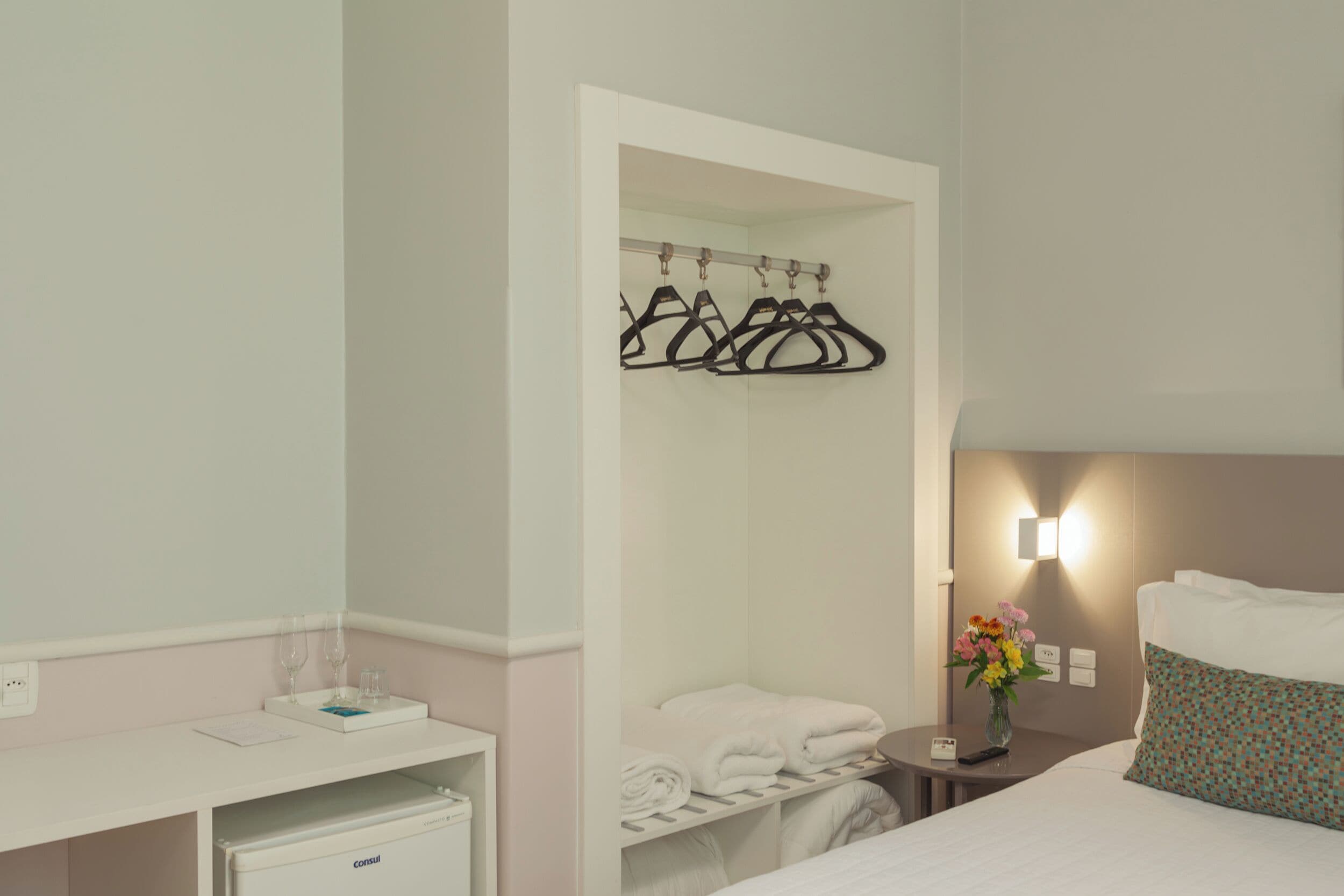 A hotel room with a bed, a bedside table with a vase of flowers, a wall-mounted light, a coat rack, and a small desk with a refrigerator.