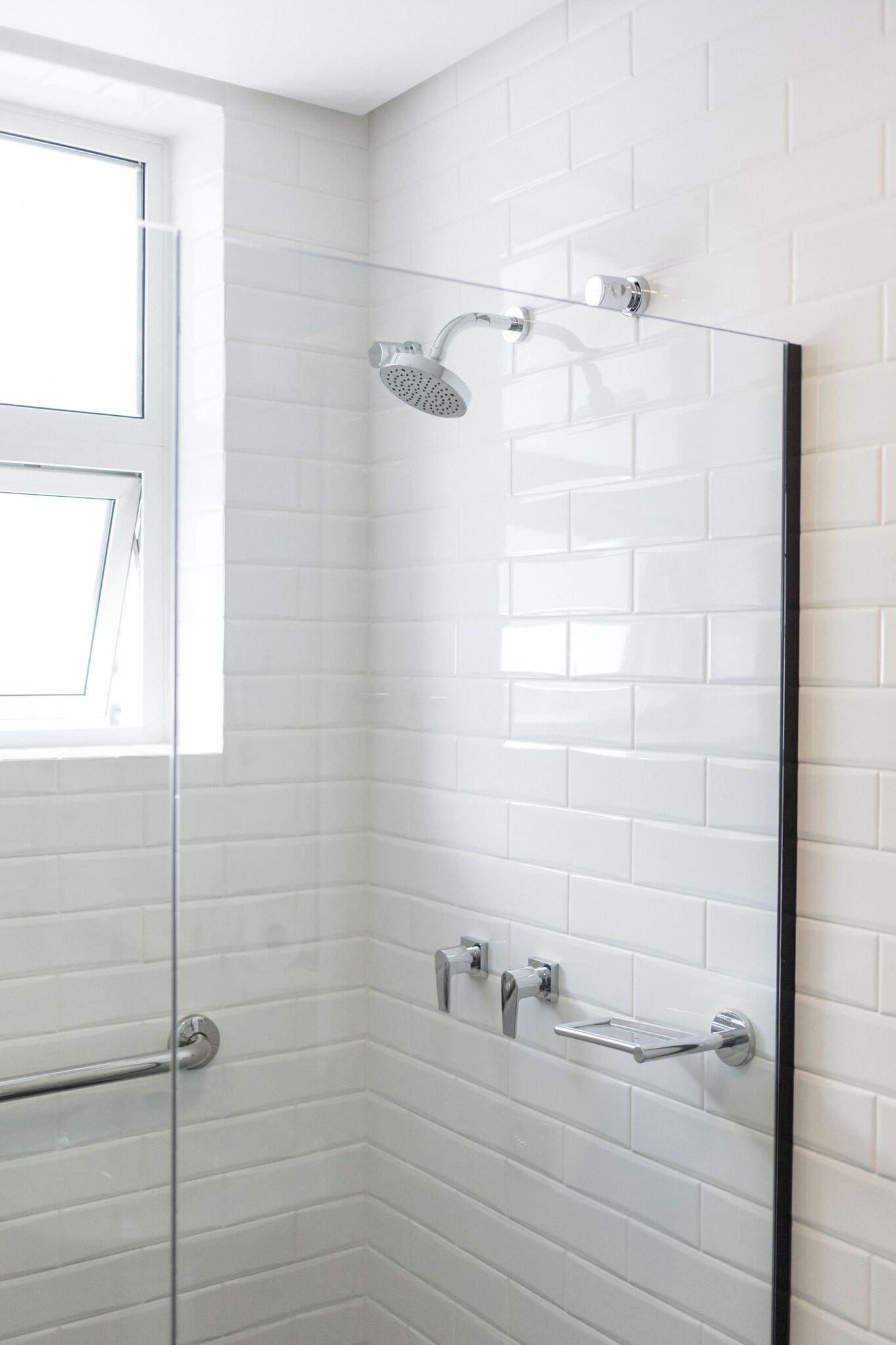 Un baño moderno con azulejos blancos tipo metro, cabina de ducha de vidrio y ventana.