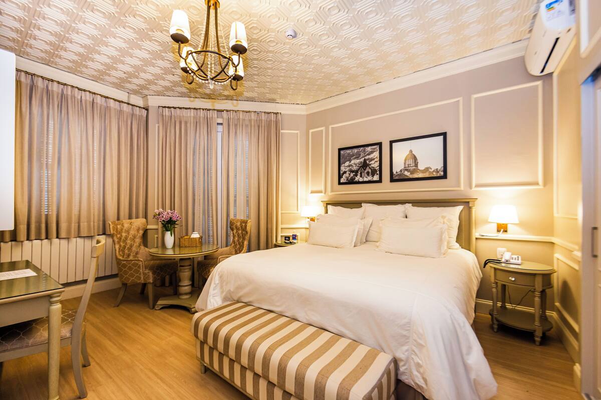 A hotel room with a large bed, a desk, a chair, a small table with a vase, and a bench.