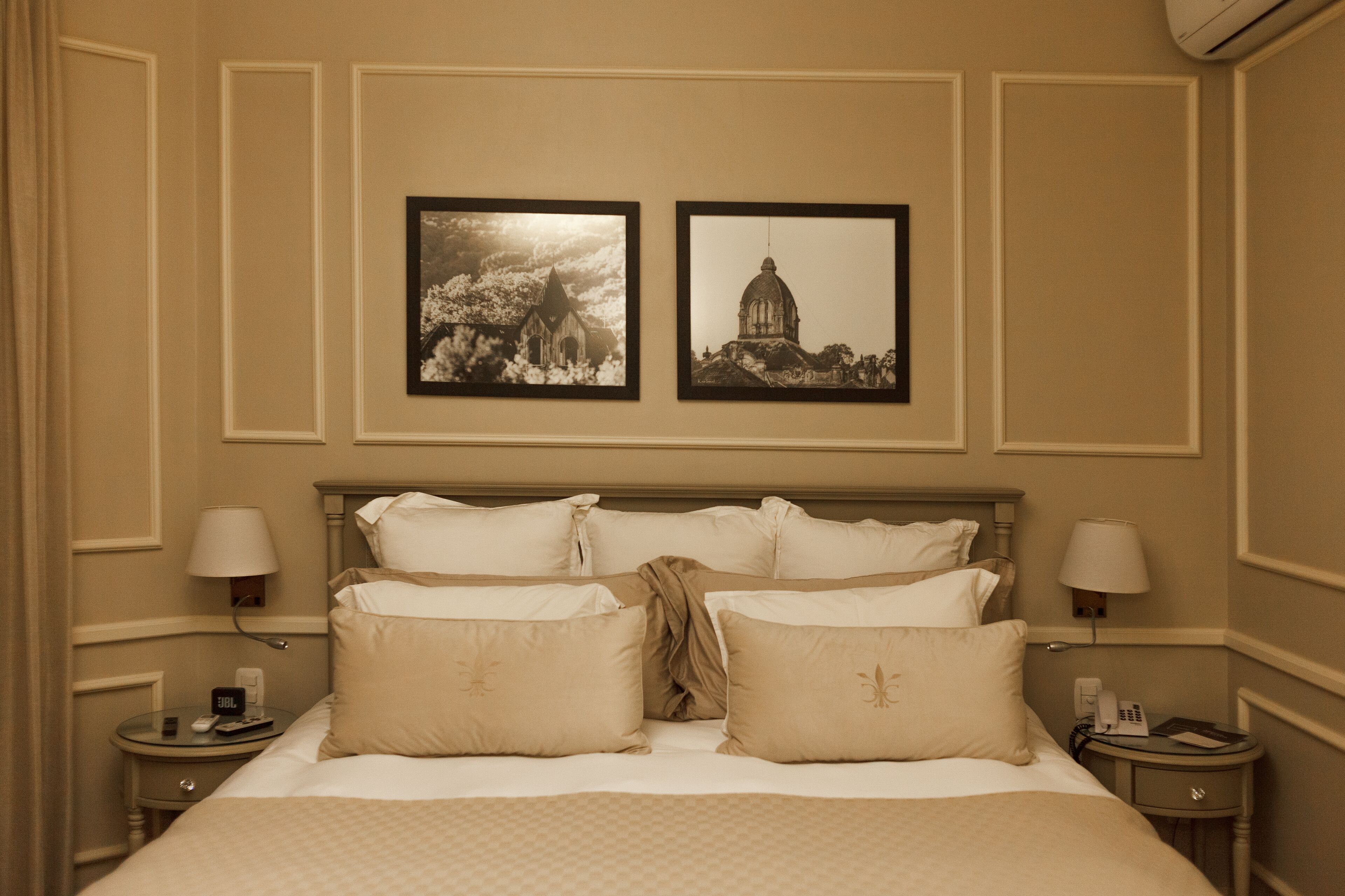 Una habitación de hotel con una cama grande, dos mesitas de noche, una lámpara de pared y dos cuadros enmarcados sobre la cama.