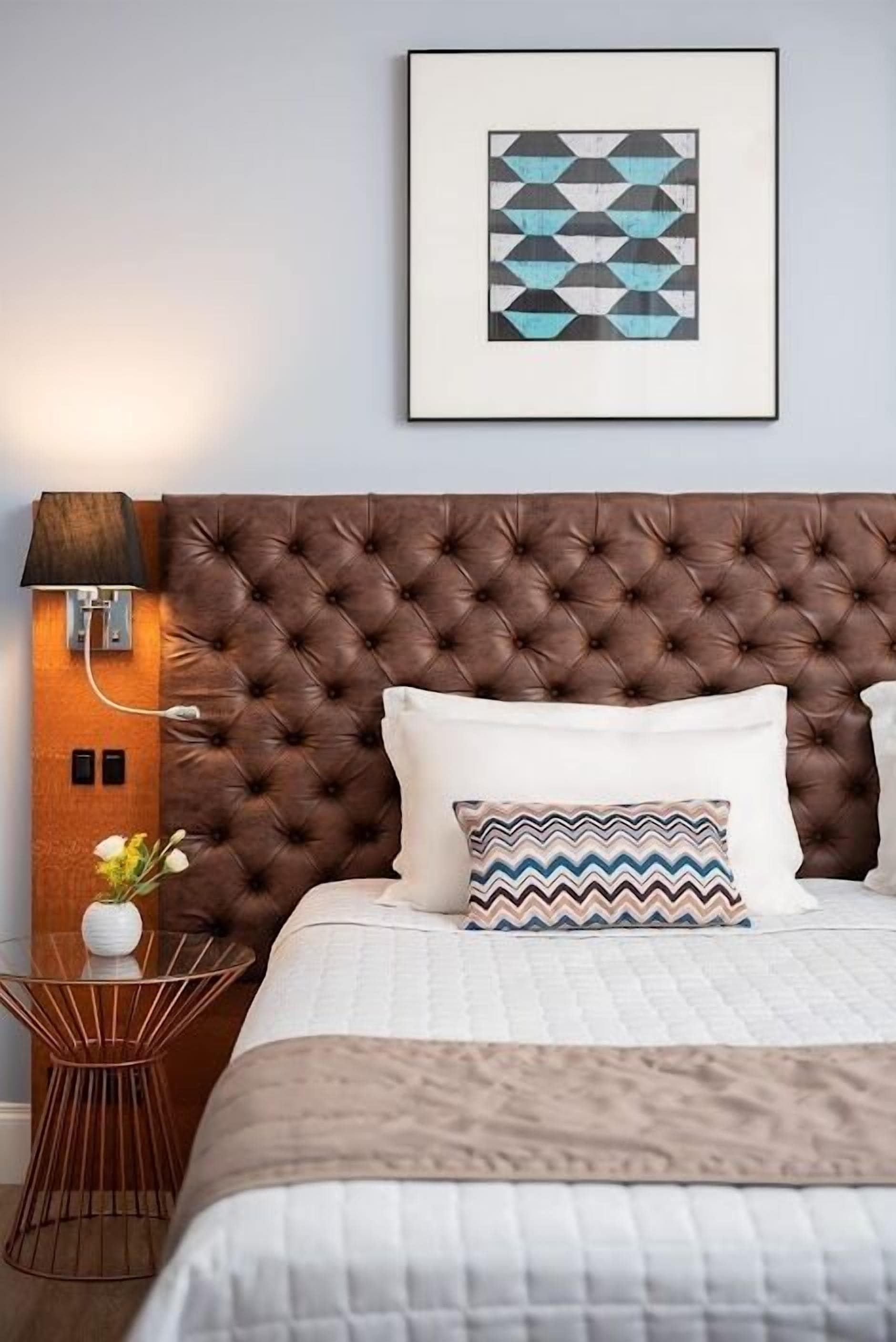 Un dormitorio con cabecera capitonada, una mesita de noche con lámpara, una obra de arte abstracta enmarcada y un cojín estampado.