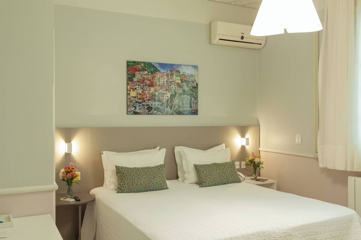 A hotel room with a bed, two bedside tables, a nightstand, a wall-mounted air conditioner, and a painting of a coastal village.