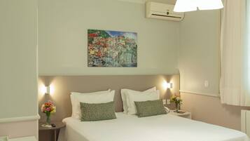 A hotel room with a bed, two bedside tables, a nightstand, a wall-mounted air conditioner, and a painting of a coastal village.