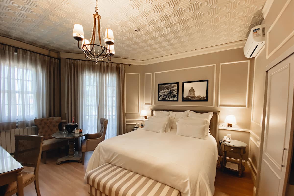 A hotel room with a large bed, a desk, a chair, a bench, a chandelier, and two wall-mounted lamps.