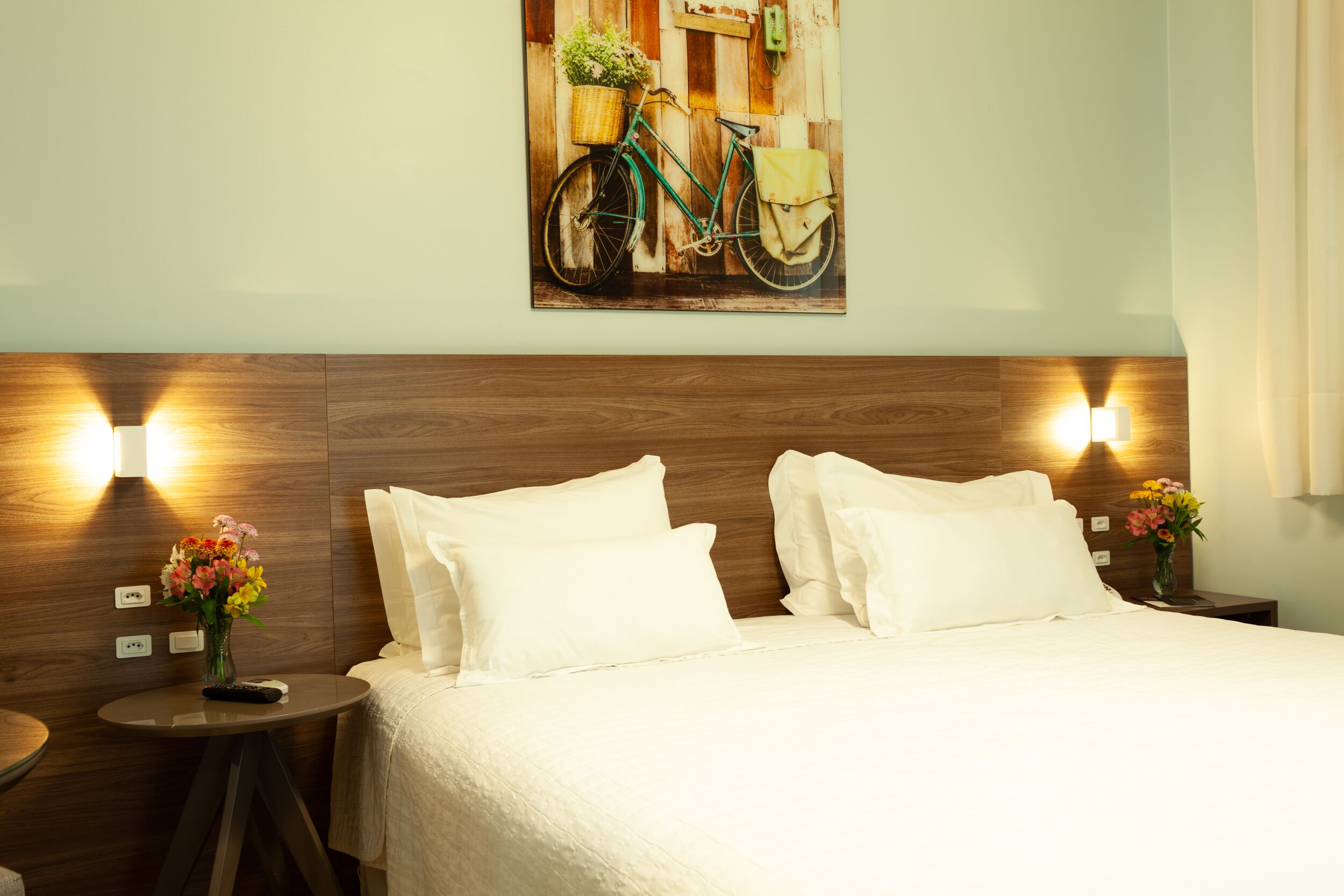 A hotel room with a wooden headboard, two white pillows, a bed with a white bedspread, a small round table with a vase of flowers, and a painting of a bicycle on the wall.