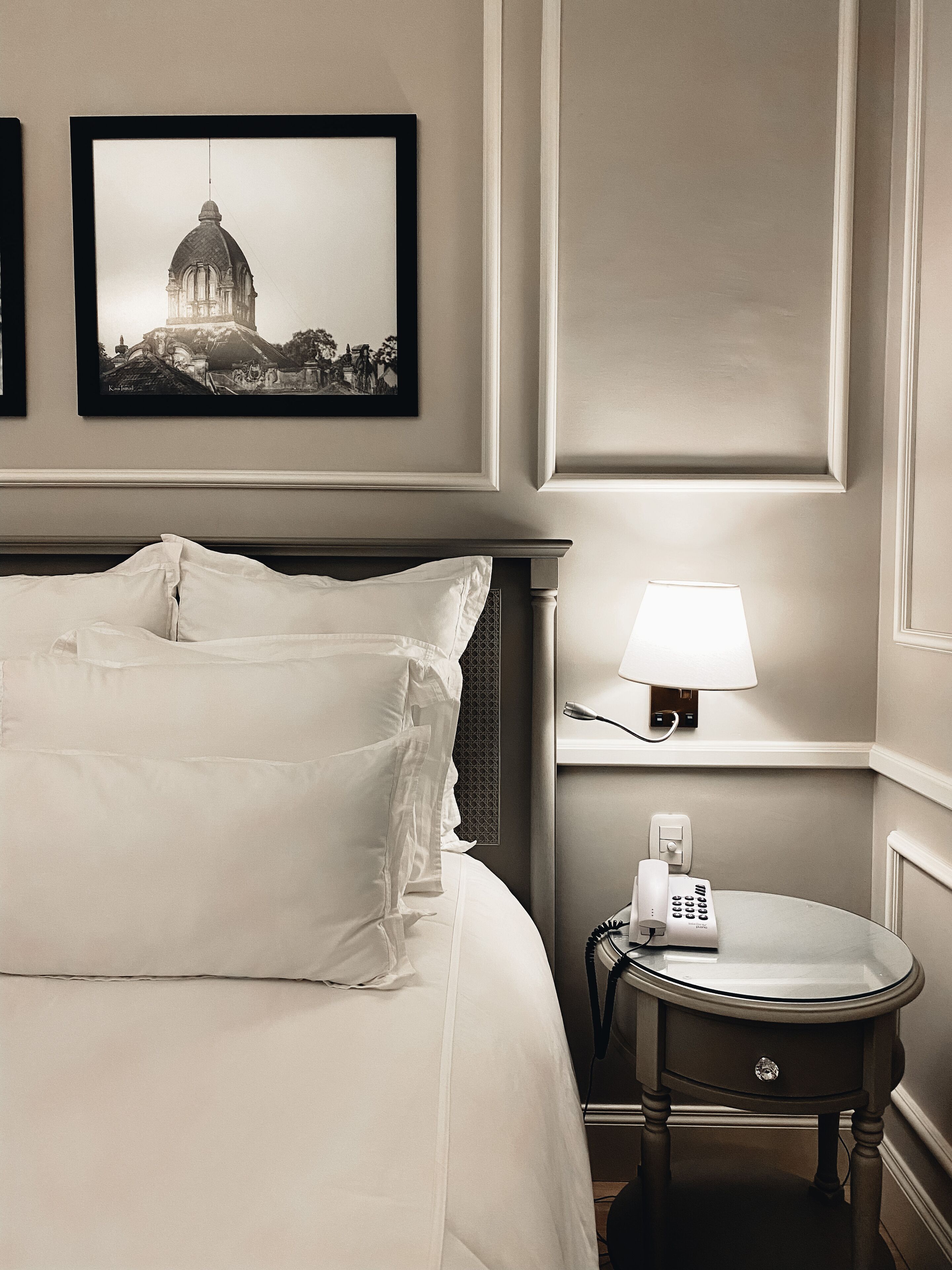 Une chambre d’hôtel avec un lit bien fait, une lampe de chevet, une petite table avec un téléphone et un tableau représentant un bâtiment accroché au mur.