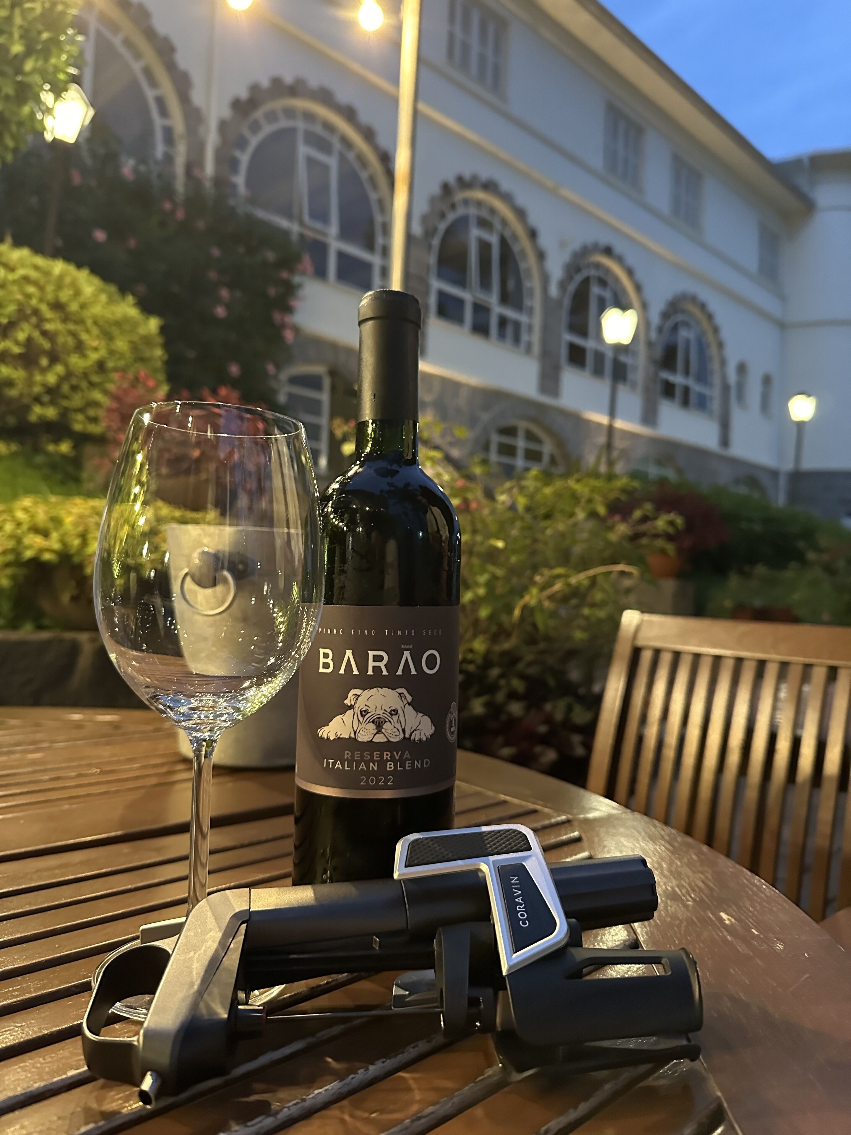A bottle of Barão Reserva Italian Blend 2022 wine and a glass on a wooden table with a bike lock and a chair.