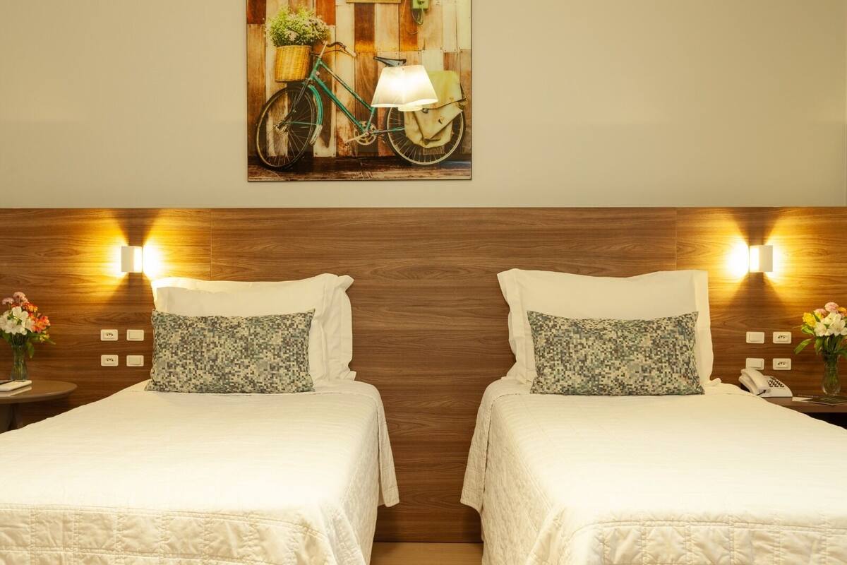 A hotel room with two beds, wooden headboard, wall-mounted lamps, and a framed picture above the beds.