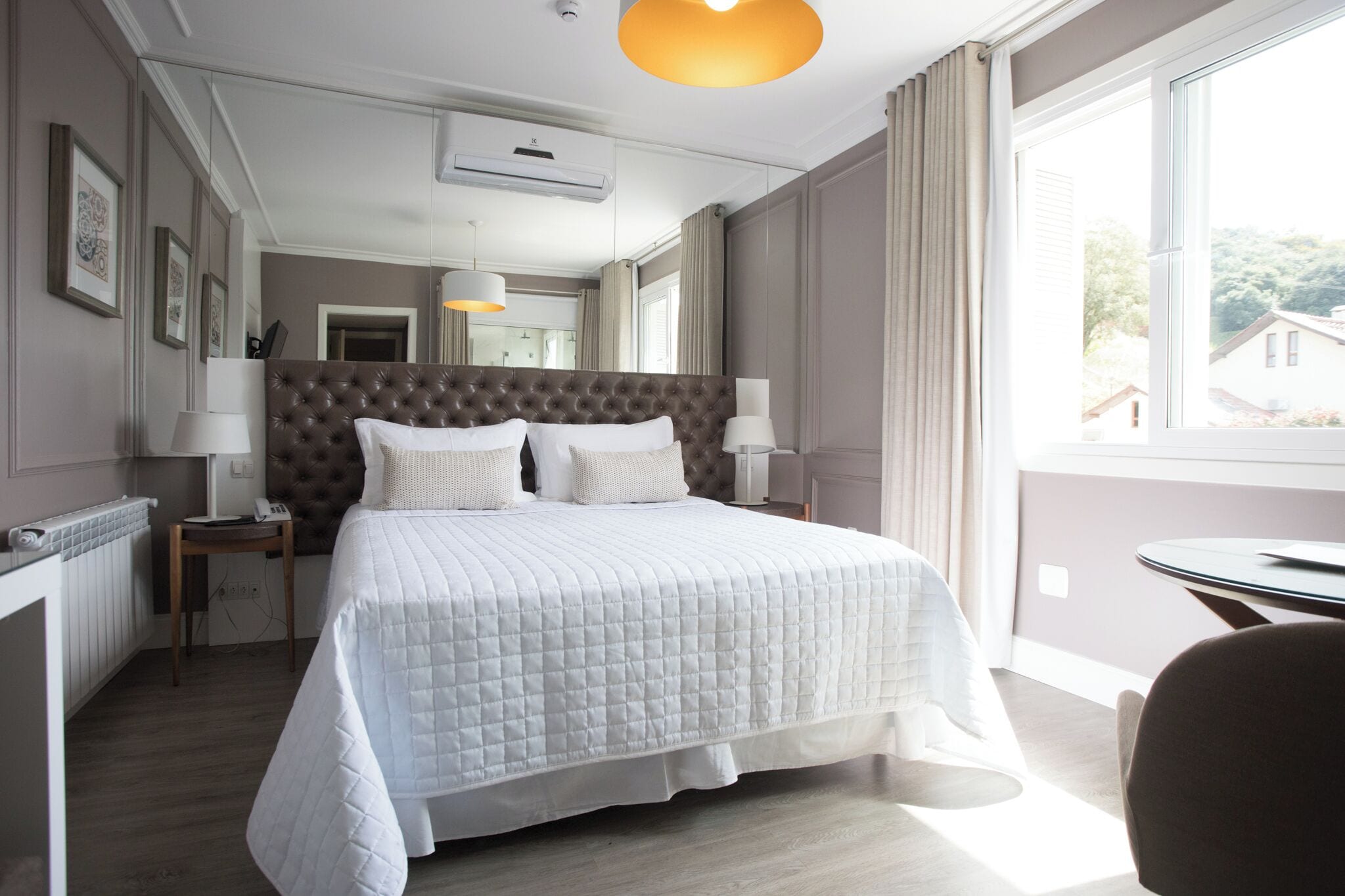 Quarto de hotel com uma cama grande, uma mesinha de cabeceira, uma cadeira, uma mesa redonda e uma janela com vista para edifícios.