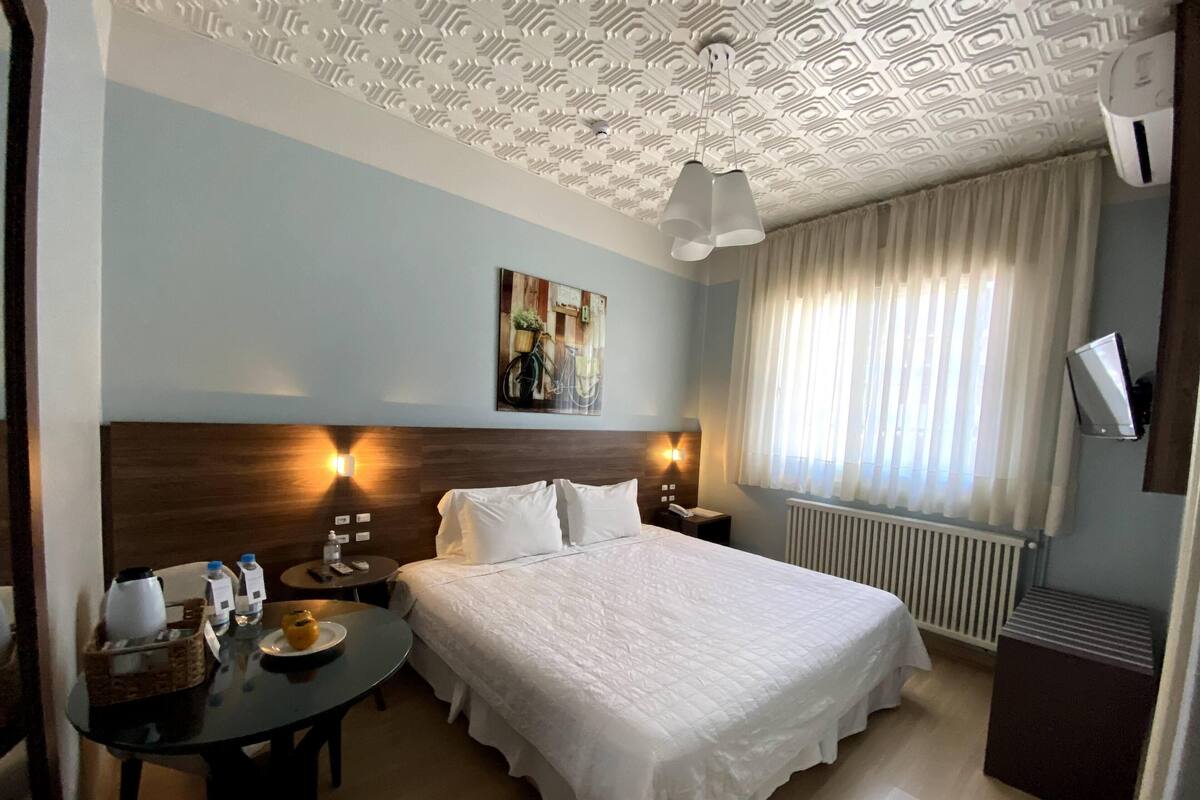 A hotel room with a large bed, a desk with a chair, a painting on the wall, and a window with curtains.