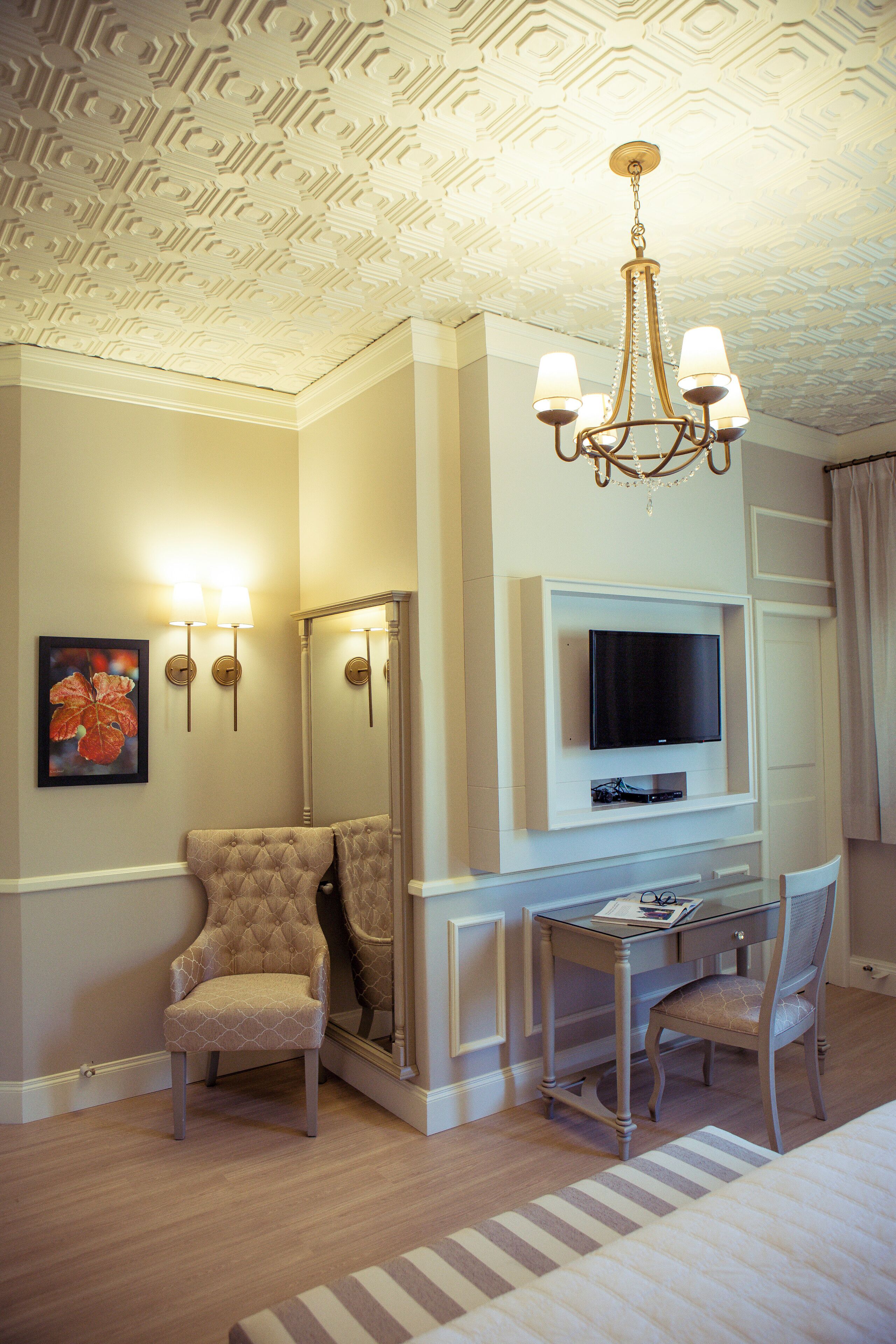 A hotel room with a wooden floor, a tufted armchair, a desk with a chair, a flat-screen TV, a chandelier, and a framed picture on the wall.