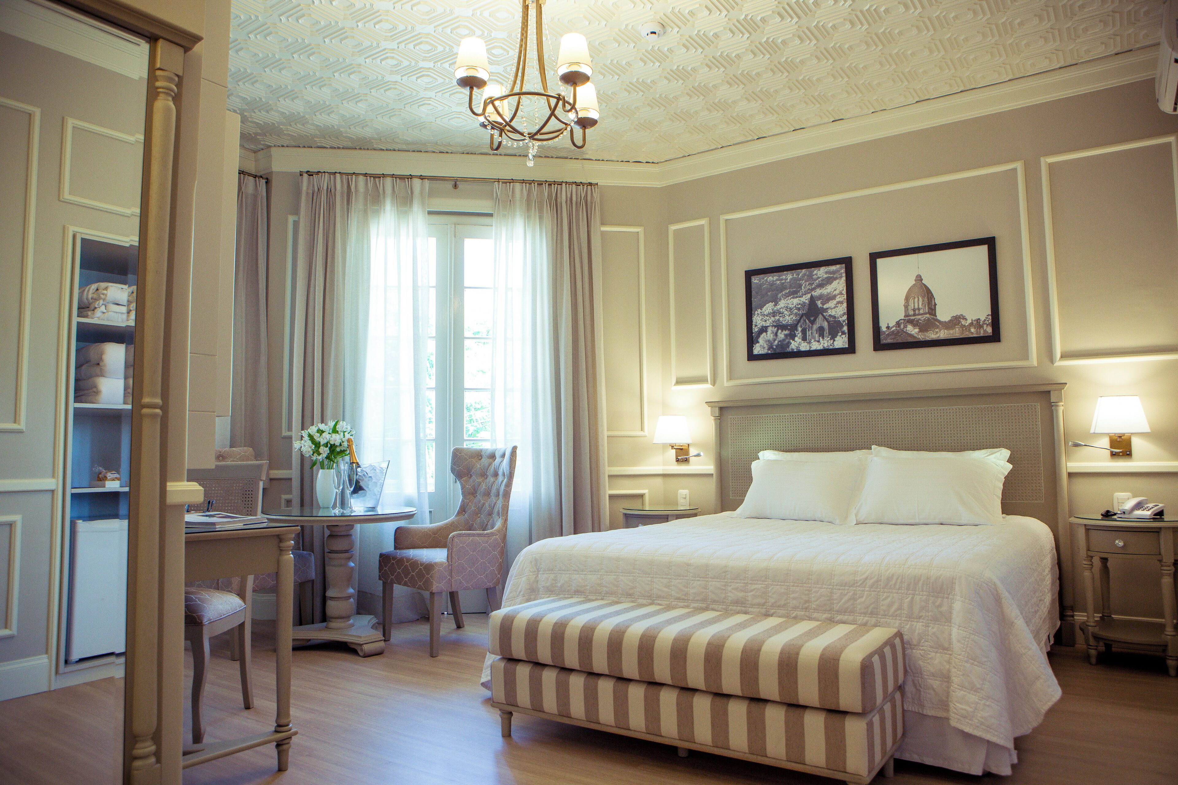 A hotel room with a large bed, a desk with a chair, a small table with a lamp, and two framed pictures on the wall.