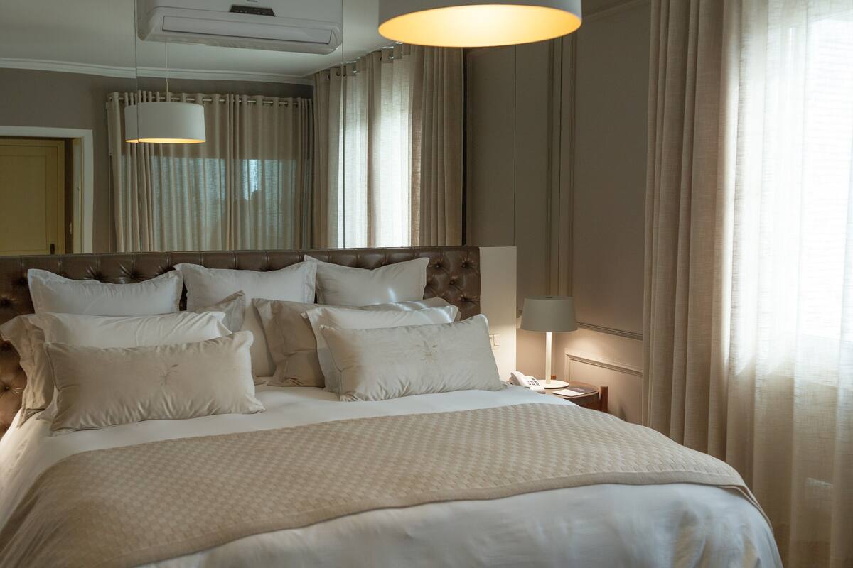 A well-appointed hotel room with a large bed, multiple pillows, a bedside lamp, and a large window with curtains.