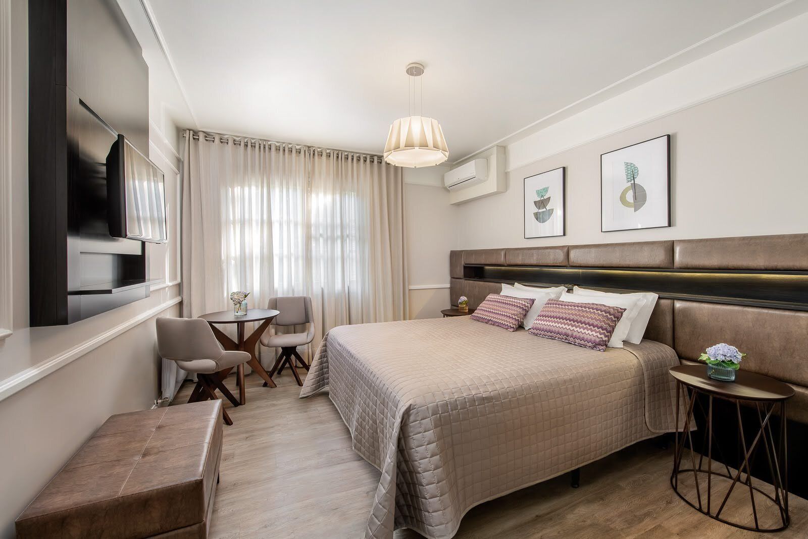 Quarto de hotel moderno com uma cama grande, uma escrivaninha, uma cadeira e uma TV fixada na parede.