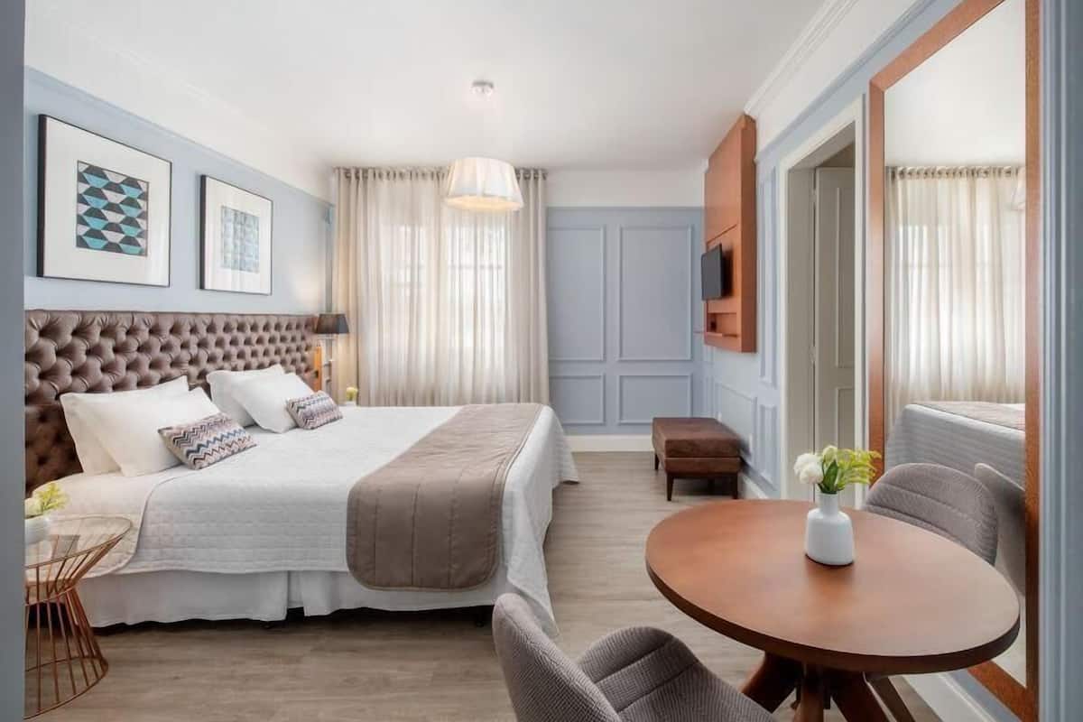 A hotel room with a large bed, a round dining table, a TV mounted on the wall, and a window with curtains.