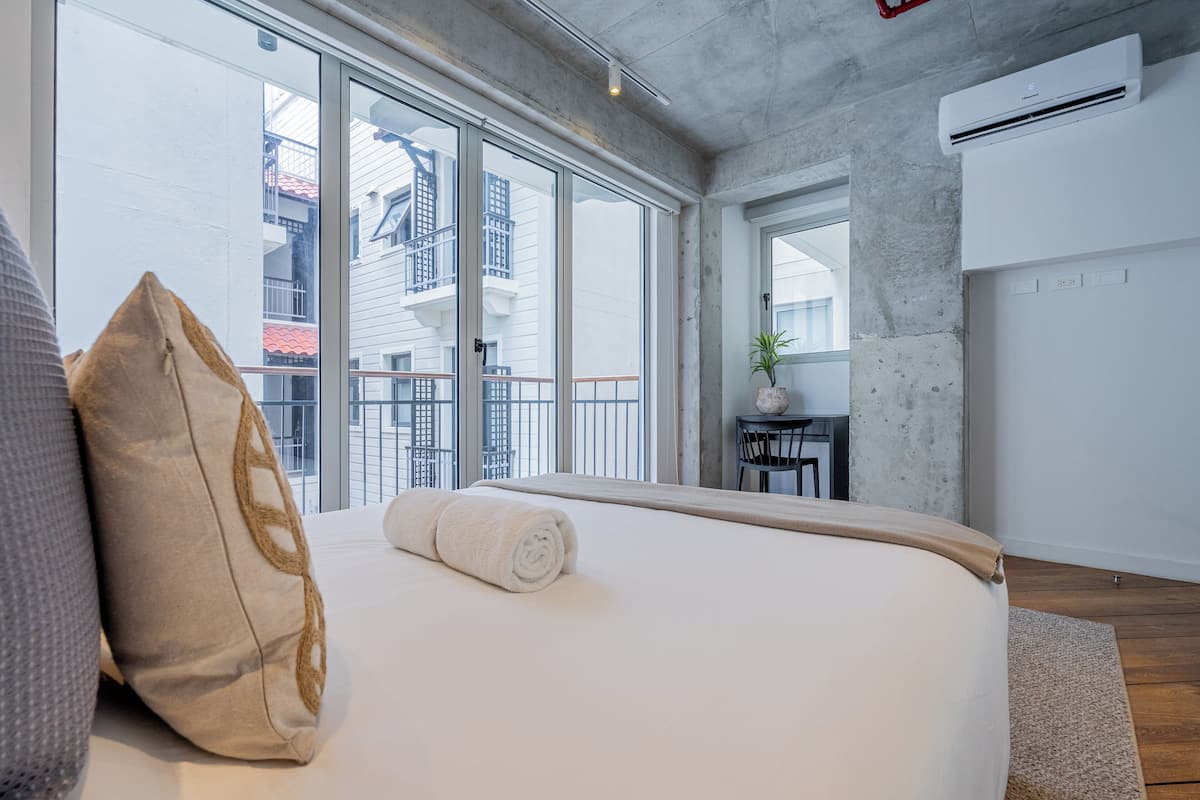 Une chambre d’hôtel moderne avec un grand lit, une vue sur la ville grâce à de grandes fenêtres du sol au plafond et un climatiseur fixé au mur.