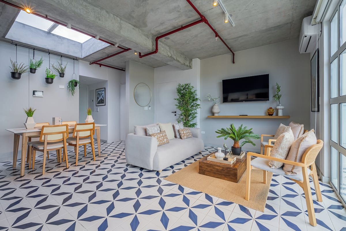 Un espace de vie moderne avec un sol à motifs géométriques, un canapé blanc, un coin repas avec des chaises en bois et diverses plantes en pot.
